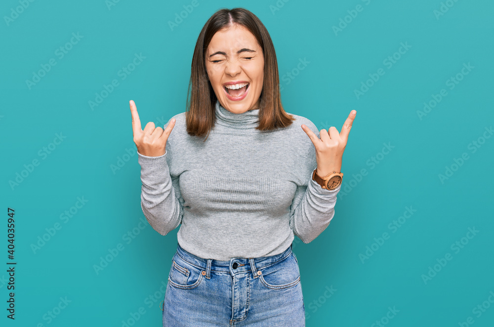 Young beautiful woman wearing casual turtleneck sweater shouting with crazy expression doing rock symbol with hands up. music star. heavy music concept. Young beautiful woman wearing casual turtleneck sweater shouting with crazy expression doing rock symbol with hands up. music star. heavy music concept.