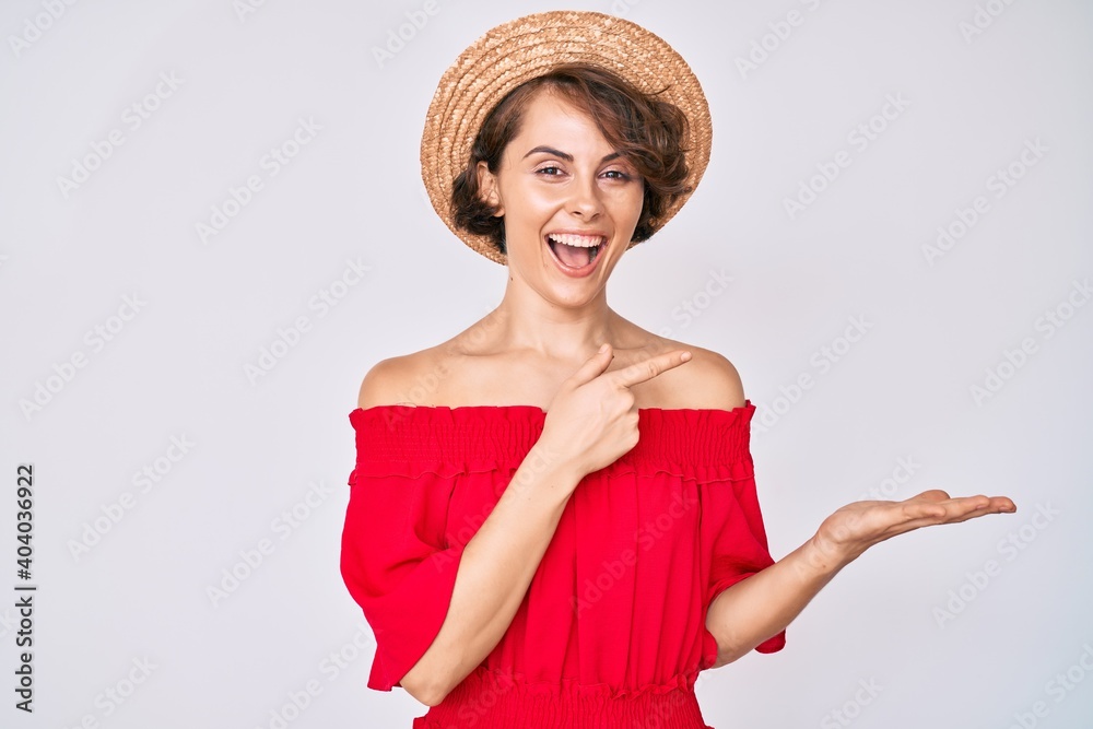 Young hispanic woman wearing summer hat amazed and smiling to the camera while presenting with hand and pointing with finger. Young hispanic woman wearing summer hat amazed and smiling to the camera while presenting with hand and pointing with finger.