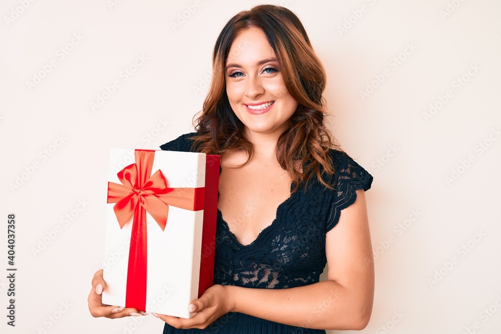 Young beautiful caucasian woman holding gift looking positive and happy standing and smiling with a confident smile showing teeth Young beautiful caucasian woman holding gift looking positive and happy standing and smiling with a confident smile showing teeth