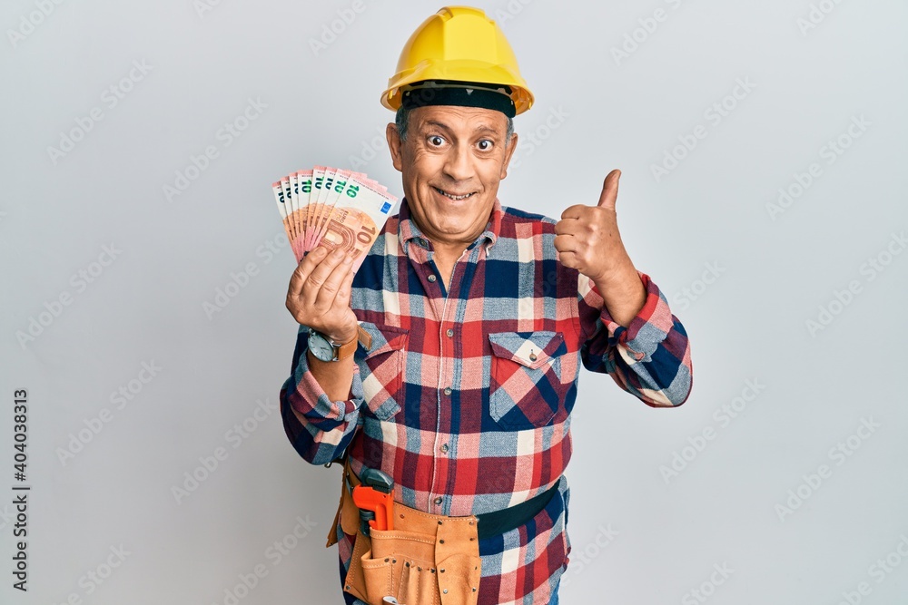 Senior hispanic man wearing handyman uniform and hardhat smiling happy and positive, thumb up doing excellent and approval sign
