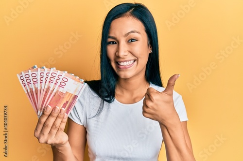 Beautiful hispanic woman holding 100 norwegian krone banknotes smiling happy and positive  thumb up doing excellent and approval sign