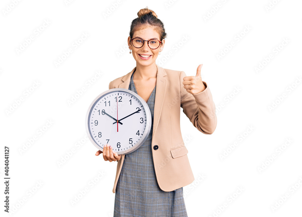 Beautiful caucasian woman with blonde hair wearing business jacket and holding clock smiling happy and positive, thumb up doing excellent and approval sign Beautiful caucasian woman with blonde hair wearing business jacket and holding clock smiling happy and positive, thumb up doing excellent and approval sign
