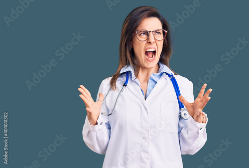 Young beautiful woman wearing doctor stethoscope and glasses crazy and mad shouting and yelling with aggressive expression and arms raised. frustration concept.
