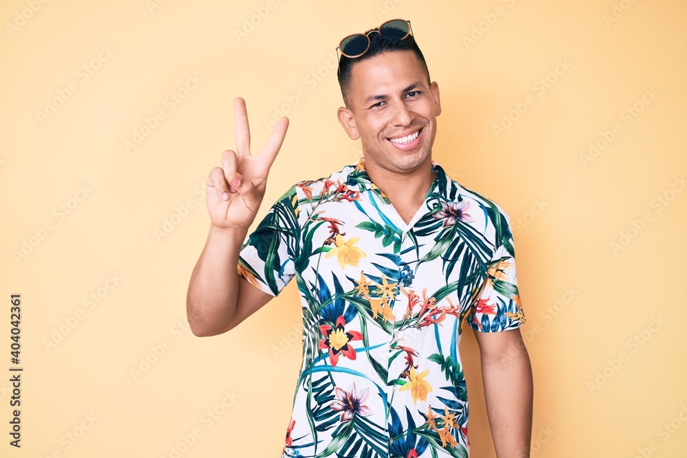 Young handsome latin man wearing summer clothes smiling with happy face winking at the camera doing victory sign. number two. Young handsome latin man wearing summer clothes smiling with happy face winking at the camera doing victory sign. number two.