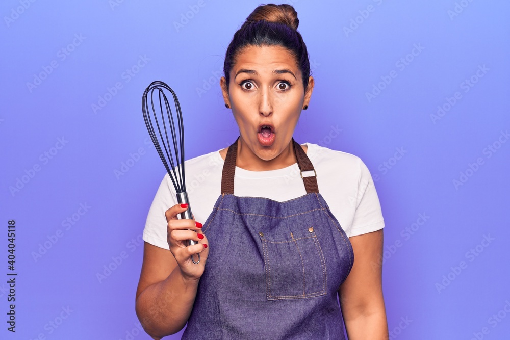 Young beautiful brunette woman wearing apron holding whisk scared and amazed with open mouth for surprise, disbelief face Young beautiful brunette woman wearing apron holding whisk scared and amazed with open mouth for surprise, disbelief face