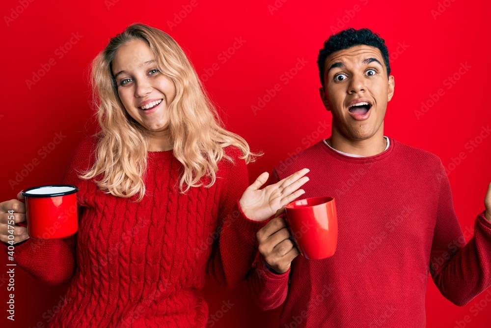 Young interracial couple holding coffee celebrating achievement with happy smile and winner expression with raised hand Young interracial couple holding coffee celebrating achievement with happy smile and winner expression with raised hand