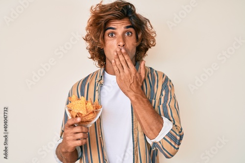 Young hispanic man holding nachos potato chips covering mouth with hand, shocked and afraid for mistake. surprised expression