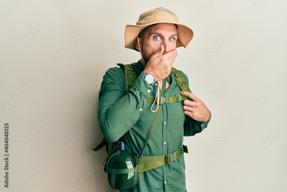 Handsome man with beard wearing explorer hat and backpack shocked covering mouth with hands for mistake. secret concept. Handsome man with beard wearing explorer hat and backpack shocked covering mouth with hands for mistake. secret concept.