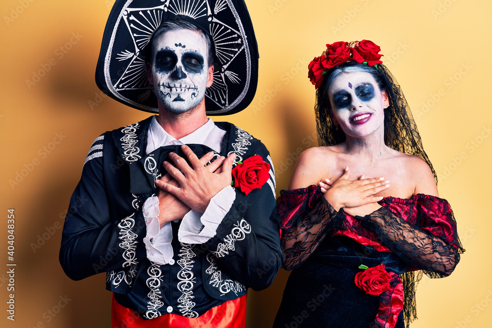 Young couple wearing mexican day of the dead costume over yellow smiling with hands on chest with closed eyes and grateful gesture on face. health concept. Young couple wearing mexican day of the dead costume over yellow smiling with hands on chest with closed eyes and grateful gesture on face. health concept.