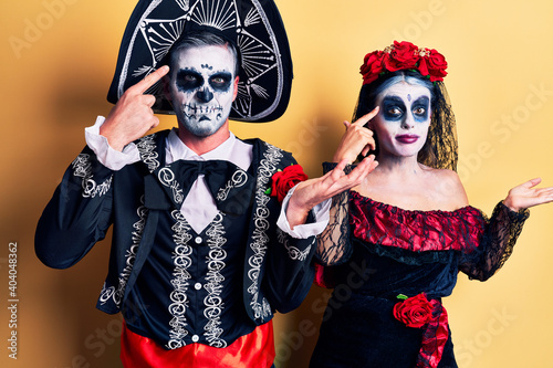 Young couple wearing mexican day of the dead costume over yellow confused and annoyed with open palm showing copy space and pointing finger to forehead. think about it.