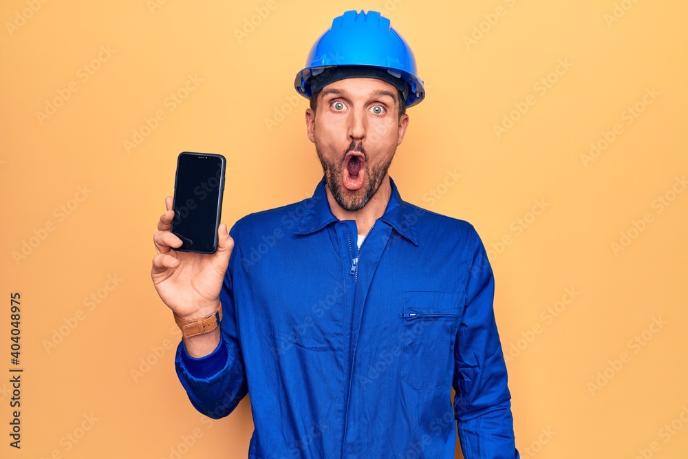 Young handsome worker man wearing uniform and hardhat holding smartphone showing screen scared and amazed with open mouth for surprise, disbelief face Young handsome worker man wearing uniform and hardhat holding smartphone showing screen scared and amazed with open mouth for surprise, disbelief face