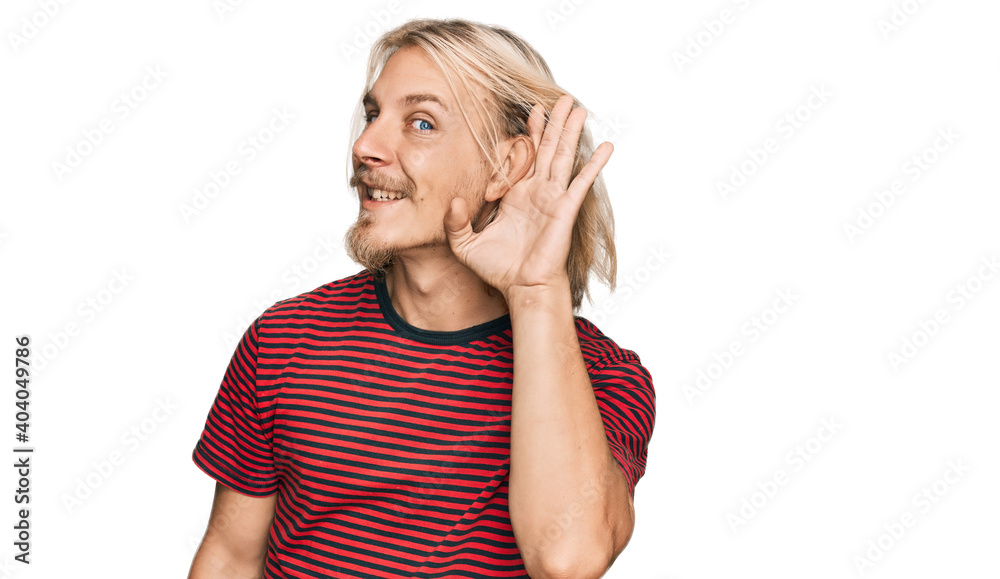 Caucasian man with blond long hair wearing casual striped t shirt smiling with hand over ear listening an hearing to rumor or gossip. deafness concept. Caucasian man with blond long hair wearing casual striped t shirt smiling with hand over ear listening an hearing to rumor or gossip. deafness concept.