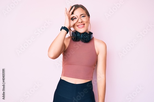 Beautiful young caucasian woman wearing gym clothes and using headphones smiling happy doing ok sign with hand on eye looking through fingers