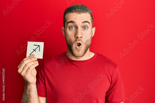 Young caucasian man holding paper with sagittarius zodiac sign scared and amazed with open mouth for surprise, disbelief face