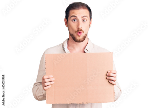 Young handsome caucasian man holding empty cardboard banner scared and amazed with open mouth for surprise, disbelief face