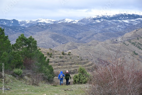 Man and woman hikers on the mountain going down. Views of pines, terraces and snow-capped mountains and windmills in the background. Cidacos Valley, La Rioja.