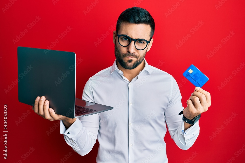 Young hispanic man wearing business style holding laptop and credit card skeptic and nervous, frowning upset because of problem. negative person. Young hispanic man wearing business style holding laptop and credit card skeptic and nervous, frowning upset because of problem. negative person.