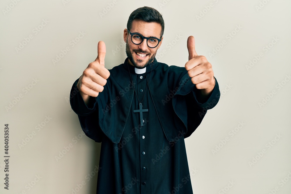 Young hispanic man wearing priest uniform standing over white background approving doing positive gesture with hand, thumbs up smiling and happy for success. winner gesture. Young hispanic man wearing priest uniform standing over white background approving doing positive gesture with hand, thumbs up smiling and happy for success. winner gesture.