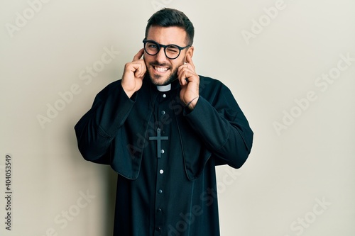 Young hispanic man wearing priest uniform standing over white background covering ears with fingers with annoyed expression for the noise of loud music. deaf concept.