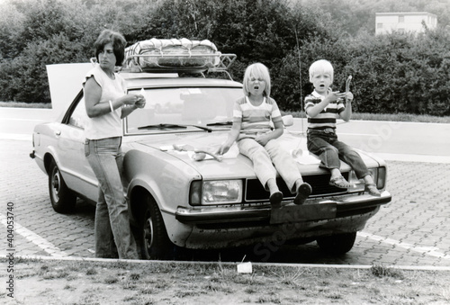 Vintage monochrome 1978 image; young mother with daugter and son sitting on hood of car eathing lunch sausages on a roadtrip holiday in Germany. 