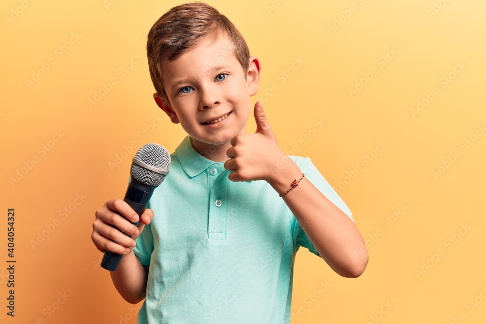 Cute blond kid singing song using microphone smiling happy and positive, thumb up doing excellent and approval sign Cute blond kid singing song using microphone smiling happy and positive, thumb up doing excellent and approval sign