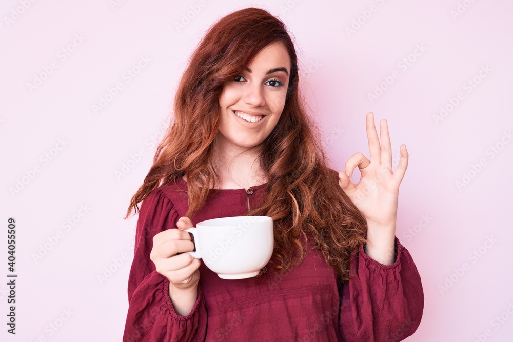 Young beautiful woman holding coffee doing ok sign with fingers, smiling friendly gesturing excellent symbol Young beautiful woman holding coffee doing ok sign with fingers, smiling friendly gesturing excellent symbol