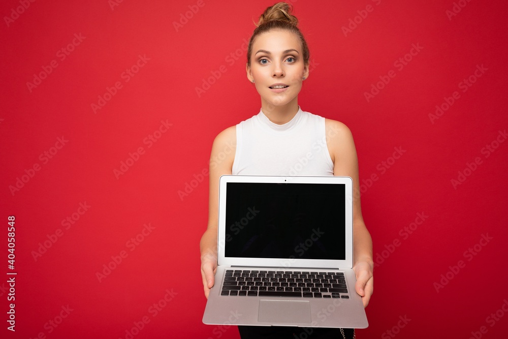 Naklejka premium Photo of beautiful blond young woman with gathered hair looking at camera holding computer laptop with empty monitor screen with mock up and copy space wearing white t-shirt isolated over red wall