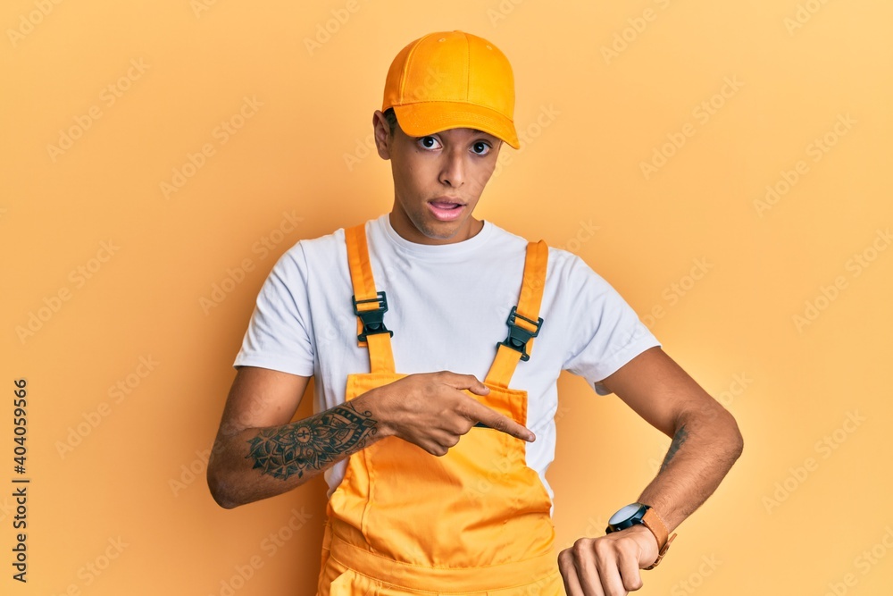 Young handsome african american man wearing handyman uniform over yellow background in hurry pointing to watch time, impatience, upset and angry for deadline delay