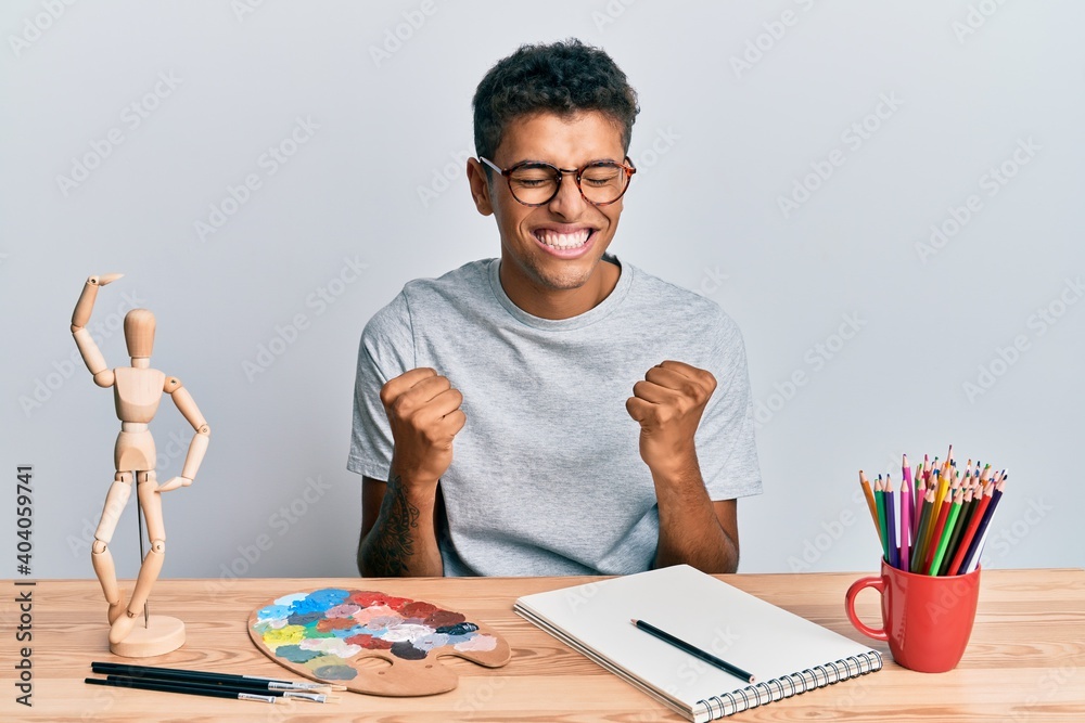 Young handsome african american man painter sitting palette and art manikin very happy and excited doing winner gesture with arms raised, smiling and screaming for success. celebration concept. Young handsome african american man painter sitting palette and art manikin very happy and excited doing winner gesture with arms raised, smiling and screaming for success. celebration concept.