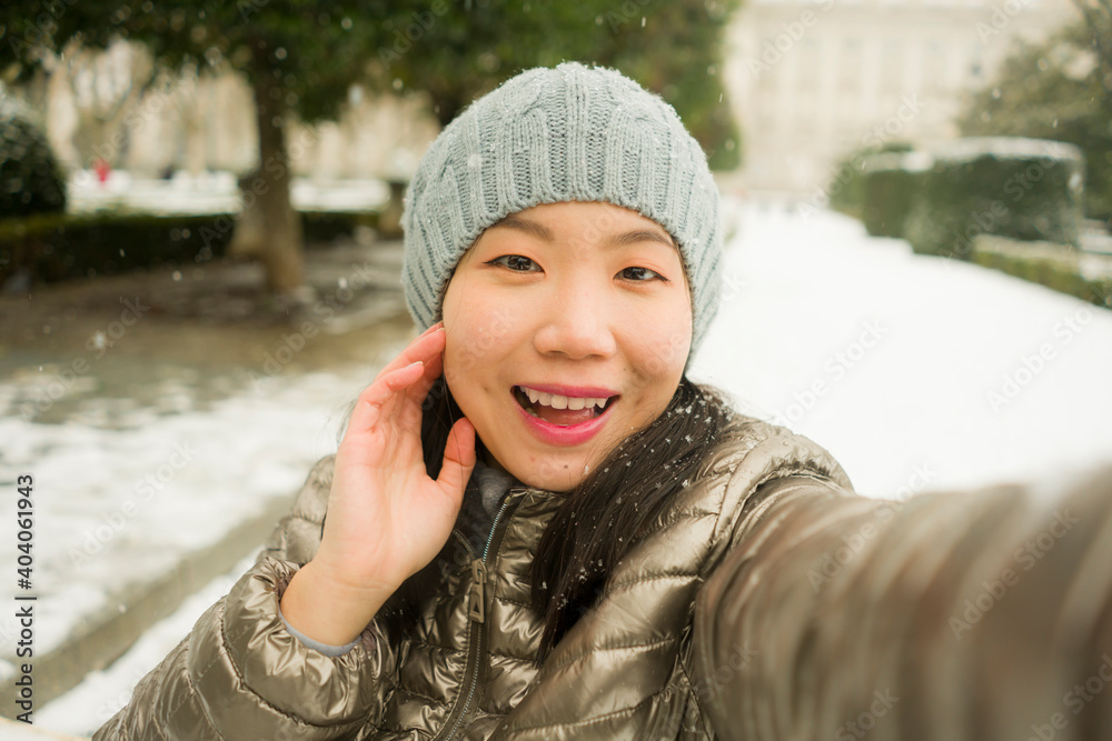 winter lifestyle portrait of young happy and beautiful Asian Korean woman taking selfie picture with mobile phone enjoying snow at city park during Christmas holidays travel