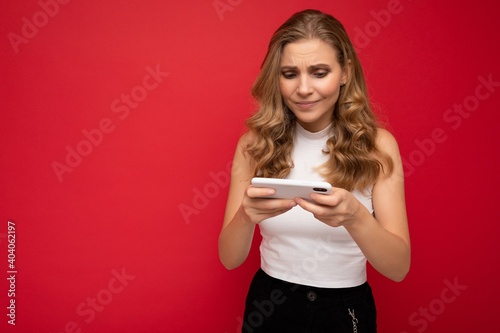 Photo of concentrated beautiful young blonde woman wearing white t-shirt isolated on red background using smartphone and playing phone games looking at gadjet screen photo