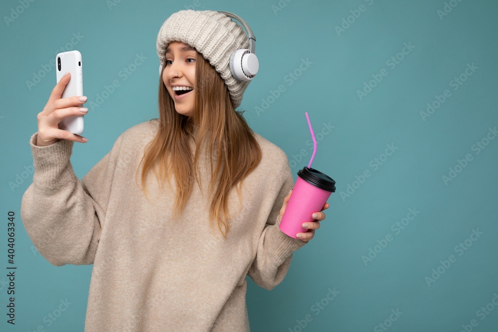 Attractive overjoyed delightful young blonde woman wearing beige sweater and beige hat white headphones isolated over blue background holding in hand and using mobile phone drinking beverage and Attractive overjoyed delightful young blonde woman wearing beige sweater and beige hat white headphones isolated over blue background holding in hand and using mobile phone drinking beverage and