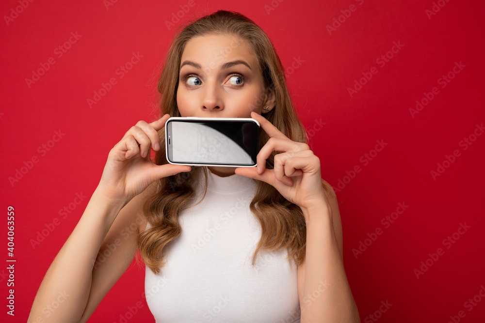attractive young blonde female person wearing white t-shirt isolated on red background with copy space holding smartphone showing phone in hand with empty screen for cutout looking to the side attractive young blonde female person wearing white t-shirt isolated on red background with copy space holding smartphone showing phone in hand with empty screen for cutout looking to the side