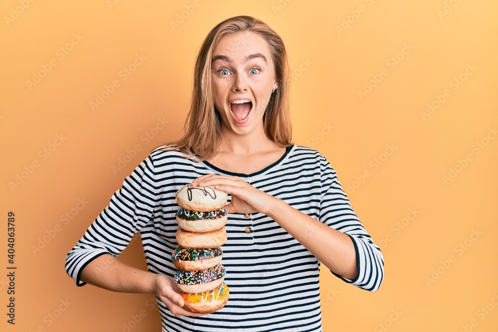 Beautiful blonde woman holding tasty colorful doughnuts celebrating crazy and amazed for success with open eyes screaming excited. Beautiful blonde woman holding tasty colorful doughnuts celebrating crazy and amazed for success with open eyes screaming excited.