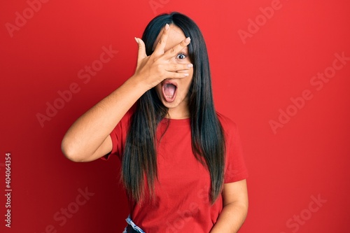 Young beautiful hispanic girl wearing casual red tshirt peeking in shock covering face and eyes with hand, looking through fingers afraid