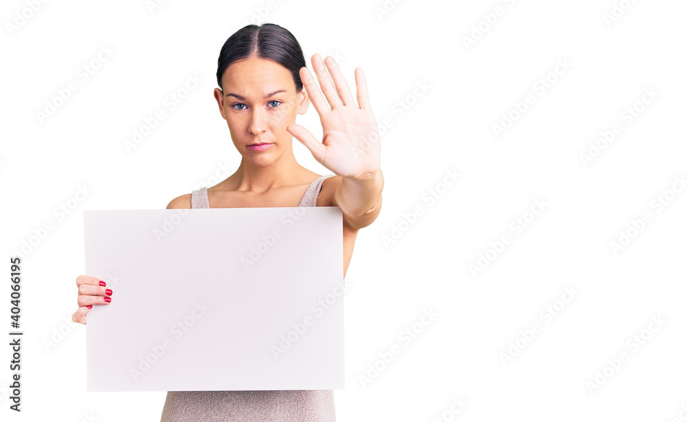 Beautiful brunette young woman holding blank empty banner with open hand doing stop sign with serious and confident expression, defense gesture Beautiful brunette young woman holding blank empty banner with open hand doing stop sign with serious and confident expression, defense gesture