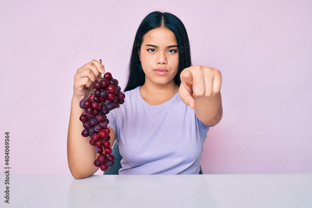 Young beautiful asian girl holding branch of fresh grapes pointing with finger to the camera and to you, confident gesture looking serious Young beautiful asian girl holding branch of fresh grapes pointing with finger to the camera and to you, confident gesture looking serious