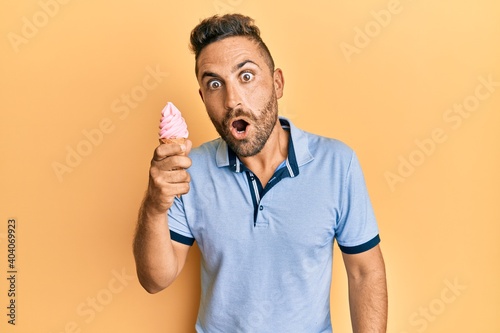 Handsome man with beard eating ice cream cone scared and amazed with open mouth for surprise, disbelief face