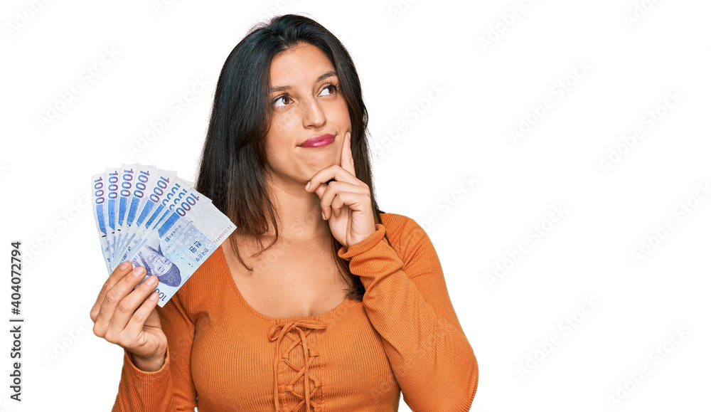 Beautiful hispanic woman holding 1000 south korean won banknotes serious face thinking about question with hand on chin, thoughtful about confusing idea Beautiful hispanic woman holding 1000 south korean won banknotes serious face thinking about question with hand on chin, thoughtful about confusing idea