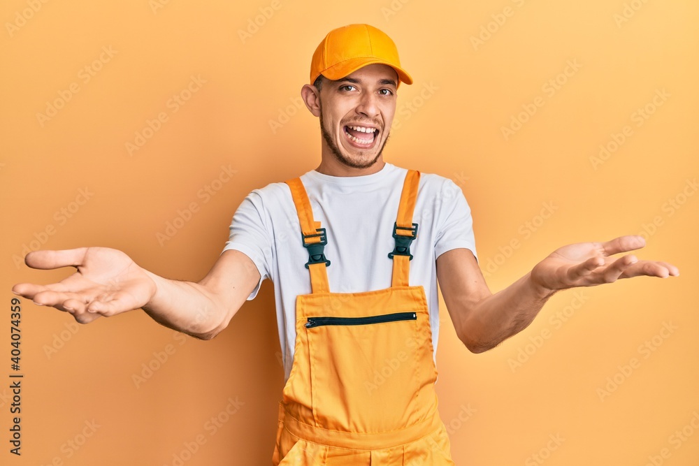 Hispanic young man wearing handyman uniform smiling cheerful offering hands giving assistance and acceptance.