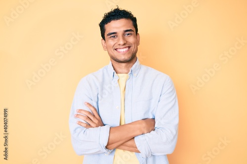Young handsome hispanic man standing over yellow background happy face smiling with crossed arms looking at the camera. positive person.