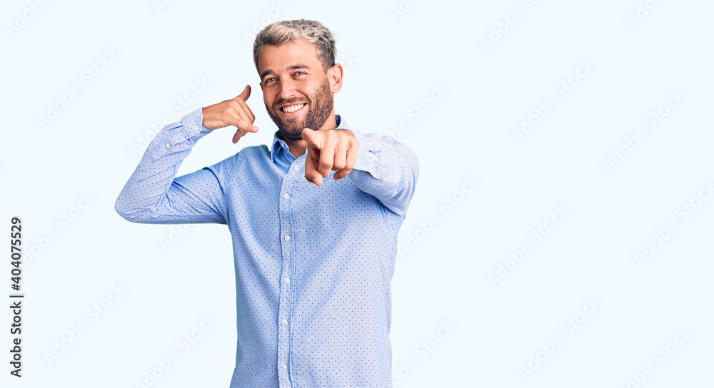 Young handsome blond man wearing elegant shirt smiling doing talking on the telephone gesture and pointing to you. call me.