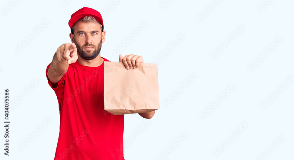 Naklejka premium Young handsome blond man holding delivery paper bag pointing with finger to the camera and to you, confident gesture looking serious
