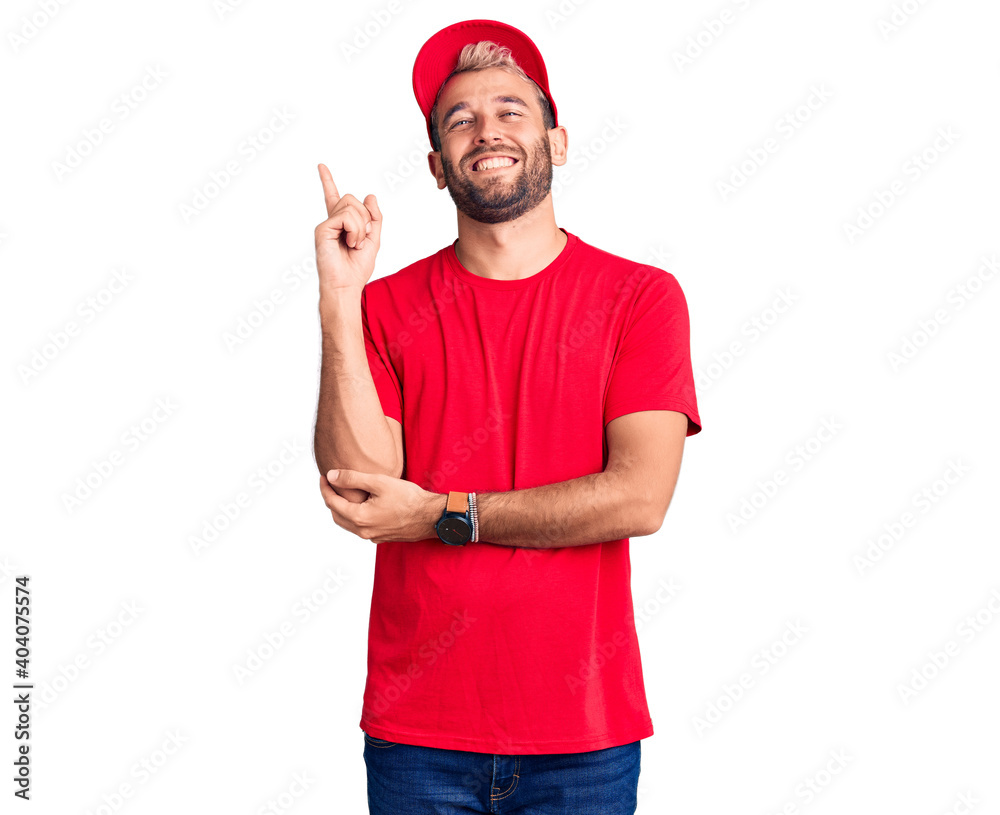Naklejka premium Young handsome blond man wearing t-shirt and cap with a big smile on face, pointing with hand and finger to the side looking at the camera.