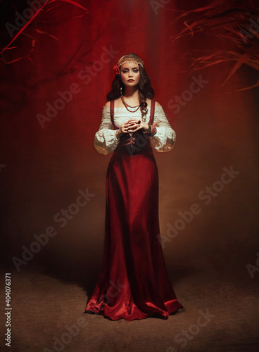 Young beautiful gypsy woman stands in a dark room. Long black flowing hair, rose hairpin. Red ethnic vintage dress, fortune teller costume. Gold jewelry. Mystical fantasy girl, pagan witch. Art photo photo