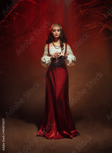 Young beautiful gypsy woman stands in a dark room. Long black flowing hair, rose hairpin. Red ethnic vintage dress, fortune teller costume. Gold jewelry. Mystical fantasy girl, pagan witch. Art photo