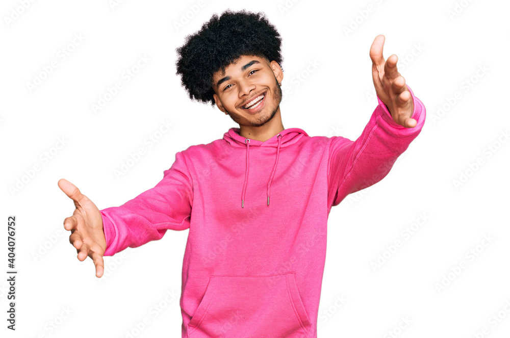 Young african american man with afro hair wearing casual pink sweatshirt looking at the camera smiling with open arms for hug. cheerful expression embracing happiness. Young african american man with afro hair wearing casual pink sweatshirt looking at the camera smiling with open arms for hug. cheerful expression embracing happiness.
