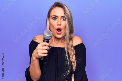 Young beautiful blonde singer woman singing using microphone over purple background scared and amazed with open mouth for surprise, disbelief face
