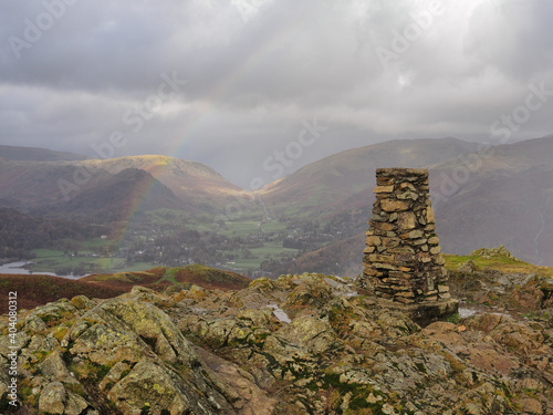 Triangulation point at the top of Loughrigg Fell overlooking Grasmere with a rainbow arcing across the fells under a stormy sky, Lake District, UK photo