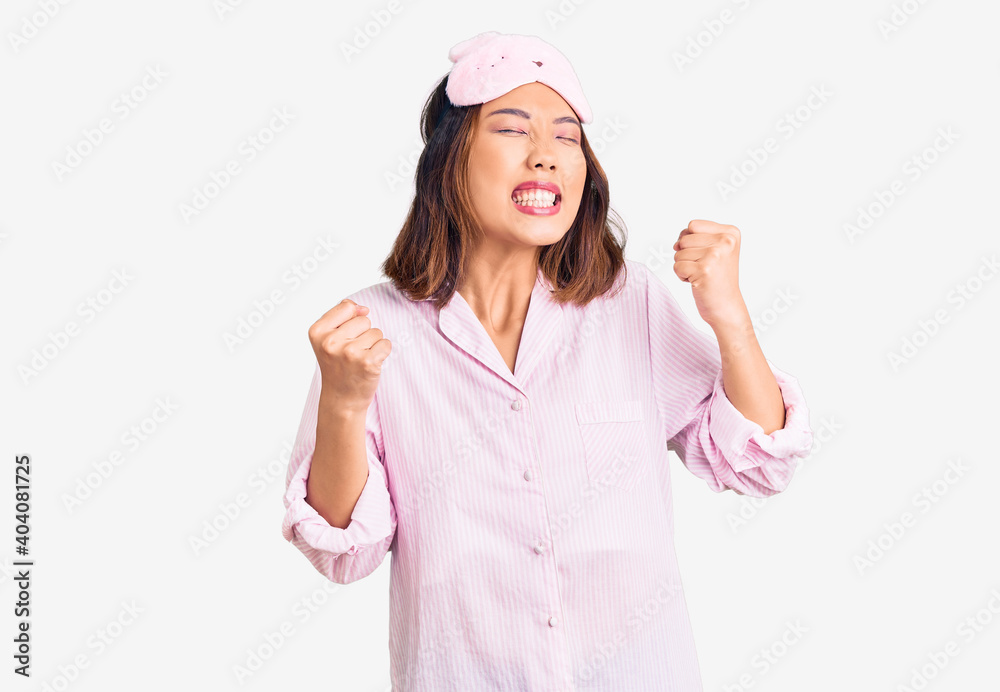Young beautiful chinese girl wearing sleep mask and pajama excited for success with arms raised and eyes closed celebrating victory smiling. winner concept. Young beautiful chinese girl wearing sleep mask and pajama excited for success with arms raised and eyes closed celebrating victory smiling. winner concept.